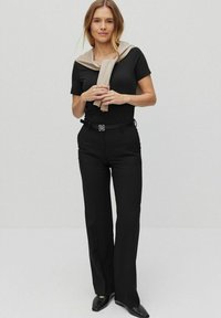 Nainen seisoo yllään musta lyhythihainen paita, mustat housut, joissa on koristevyö, mustat kengät ja beige villatakki hartioilla.