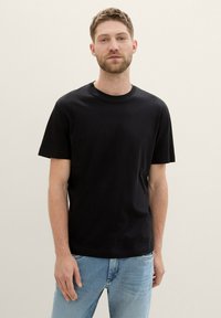 Tricou negru cu mânecă scurtă, dintr-un material neted, cu gât rotund, purtat cu blugi albaștri deschiși. Design simplu, fără modele sau accente.