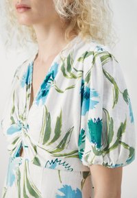 Femme portant une robe blanche avec un imprimé floral bleu et vert et des manches courtes bouffantes sur un fond uni.