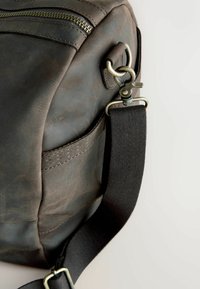 Bruine leren tas met contrasterende stiksels, een ritsvak, metalen hardware en een zwarte stoffen schouderriem. Verweerd textuur is duidelijk zichtbaar.