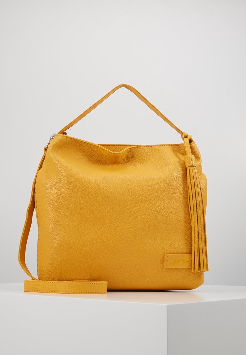 Sac à main en cuir jaune avec poignée supérieure, longue sangle réglable et pompon décoratif, posé sur une surface blanche avec un fond uni.