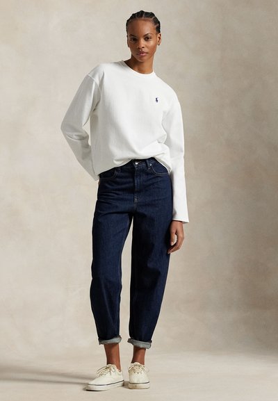 Kobieta z warkoczami, ubrana w białą bluzkę z długim rękawem, podwinięte ciemnoniebieskie dżinsy i jasnobeżowe trampki na jednolitym tle.