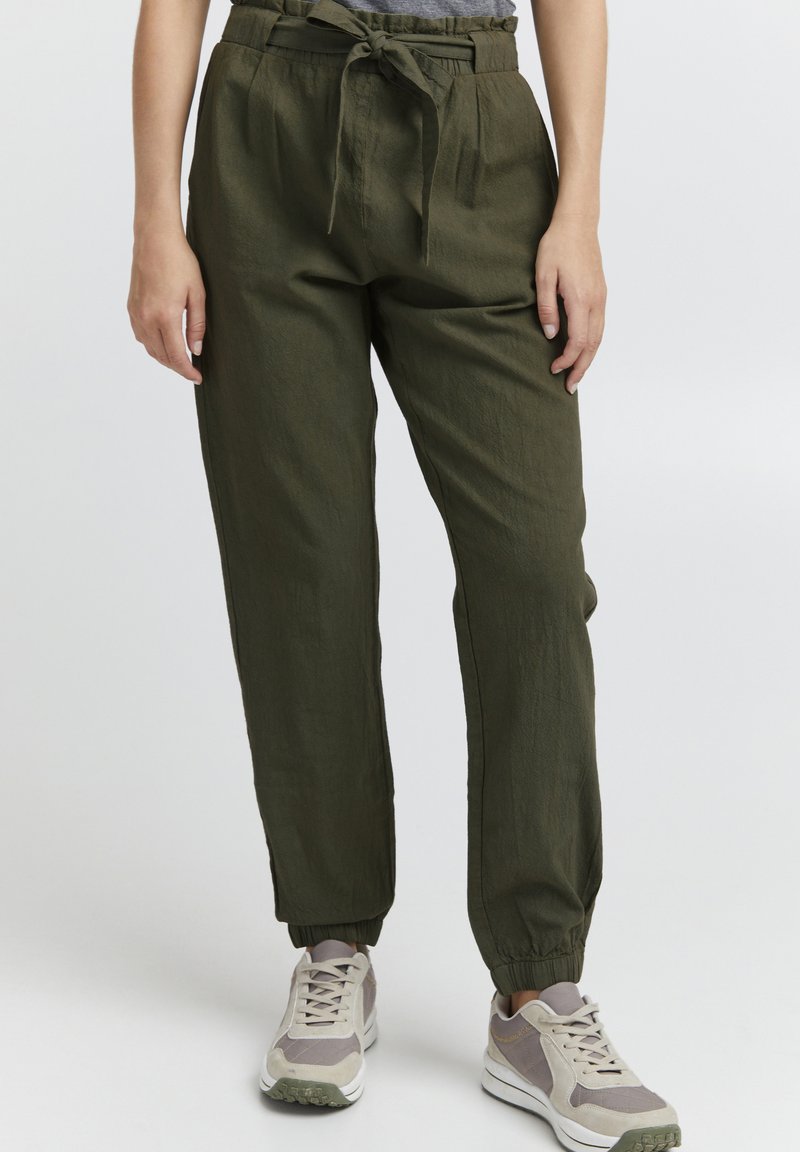 Olive green, lightweight pants with a high waist, pleated front, and drawstring tie. Cuffed ankles provide a tapered fit. Paired with gray and white sneakers.