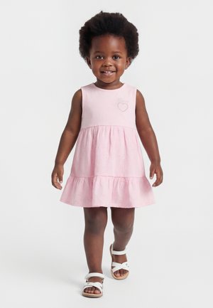 Lille pige med krøllet hår iført en ærmeløs lyserød kjole og hvide sandaler, smilende og gående fremad på en ensfarvet baggrund.