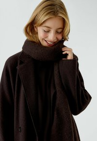Schwarzer Wolle-Mantel mit breitem Kragen, über einen dunklen gerippten Pullover und einem passenden dicken Schal getragen. Weicher, strukturierter Stoff.