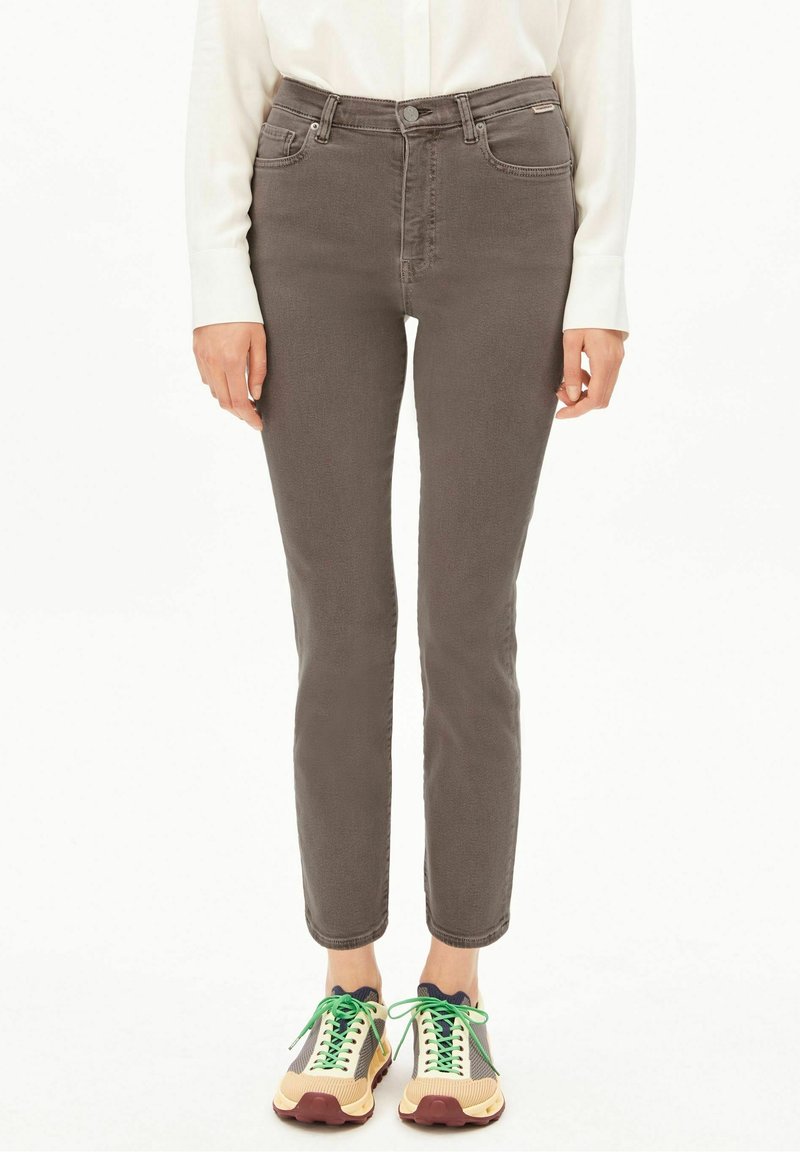 Personne portant un pantalon taupe slim, une chemise blanche à manches longues et des baskets beiges avec des lacets verts, debout sur un fond blanc.
