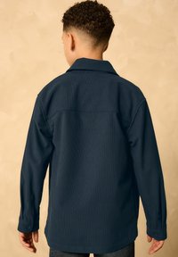 Camicia button-up di un blu scuro con maniche lunghe, tessuto testurizzato e colletto a punte. Presenta una singola yoke posteriore e polsini con bottone.