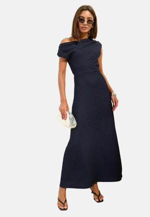 Frau trägt ein marineblaues, schulterfreies, strukturiertes Maxikleid, Sonnenbrille, schwarze Sandalen, silbernes Armband und hält eine weiße Clutch.