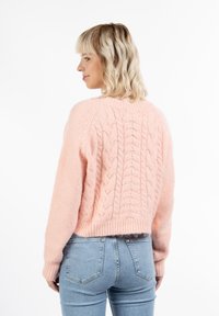 Maglione lavorato a maglia color rosa con design a treccia, maniche a raglan e orlo a coste. Indossato con jeans in denim chiaro, di texture liscia.
