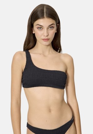 Mujer con cabello largo y castaño, vistiendo un top de bikini negro texturizado de un solo hombro y pantaletas a juego, sobre un fondo liso.