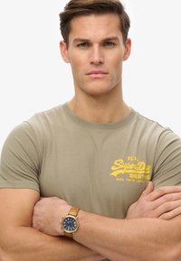 Khaki T-shirt med gult "SuperDry" logo, korte ærmer og rund halsudskæring, parret med et brunt læderrem ur med blå urskive.