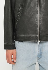 Jachetă din piele neagră cu un finisaj texturat, prezentând un fermoar frontal și o buzunare cu fermoar pe lateral. Asortată cu blugi de denim albastru deschis.