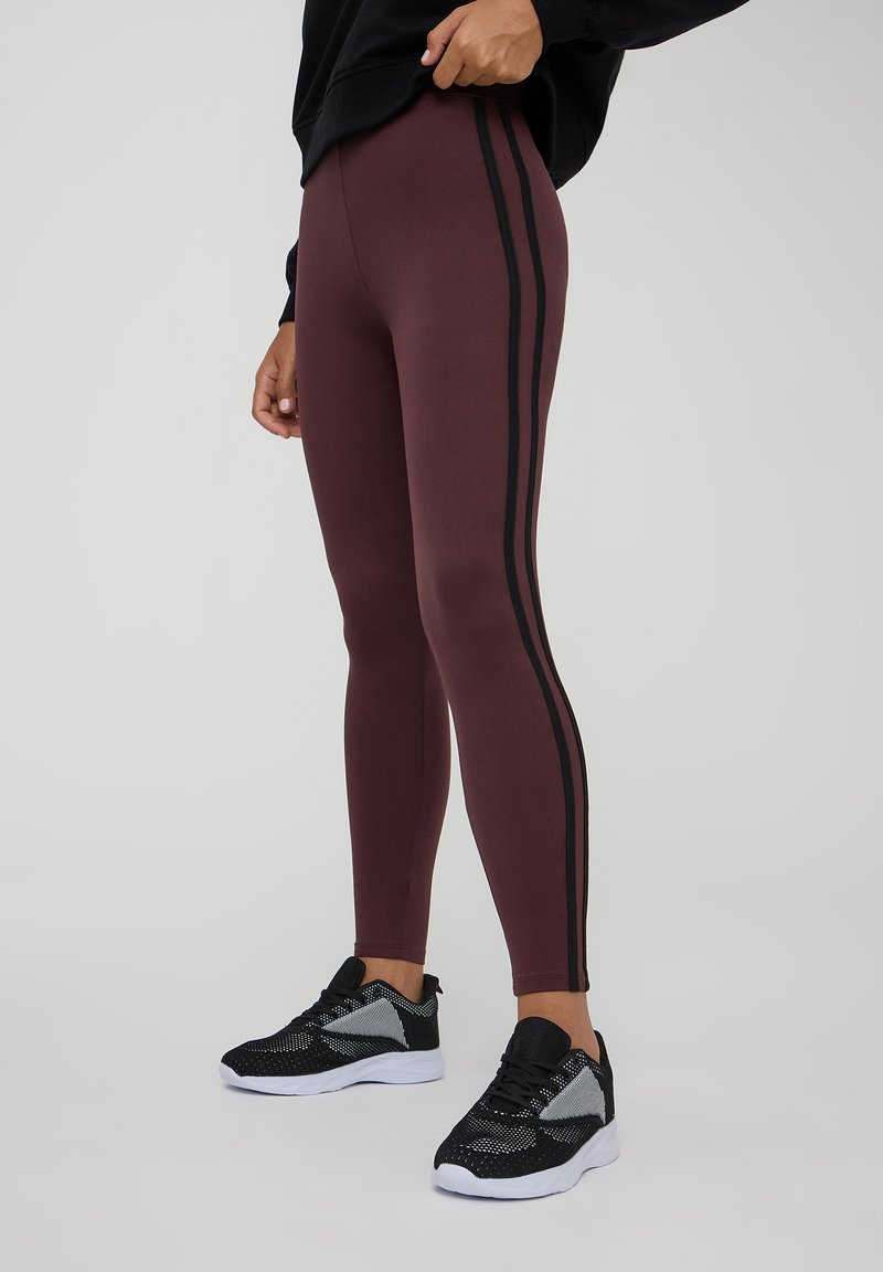 Leggings bordeaux con strisce laterali nere, realizzati in tessuto elasticizzato. Abbinati a scarpe da ginnastica nere con accenti grigi e tomaia in rete.