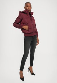 Burgunderfarget pufferjakke med hette, glidelås og ribbestrikkede mansjetter; kombinert med svarte skinny jeans med en sideaccentlinje og stiletthæler.