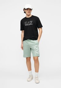 Zwart katoenen T-shirt met wit grafisch logo; muntgroene katoenen korte broek met zijlogo; witte sneakers; witte pet; neutrale achtergrond.
