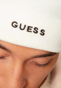 Beanie bianco lavorato a maglia con logo "GUESS" ricamato in nero, caratterizzato da una texture a coste e una vestibilità aderente. Ingrandimento del cappello sopra la fronte di una persona.