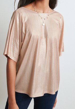 Vrouw die een glanzende, lichtroze blouse met korte mouwen draagt, met gelaagde gouden kettingen en donkerblauwe spijkerbroek.