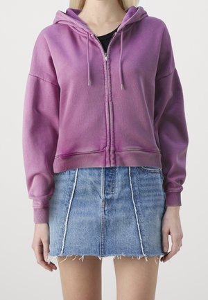 Frau trägt einen violetten Zip-Hoodie und einen blauen Jeans-Minirock mit ausgefranstem Saum und steht vor einem schlichten hellen Hintergrund.