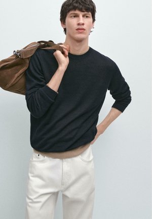 Jeune homme portant un pull noir et un pantalon blanc, tenant un sac marron sur l'épaule, debout devant un fond uni gris clair.