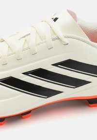 Botín de fútbol blanco con rayas negras y acentos naranjas, con material sintético texturizado, cordones planos y un diseño ligero.