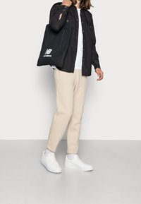 Czarna koszula na guziki, biała koszulka, beżowe spodnie typu tapered, białe sneakersy oraz czarna torba na ramię z logo "New Balance".