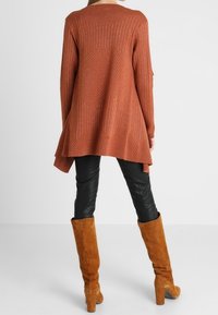 Rust-colored long-sleeve tunic with a textured pattern, flared hem; paired with tight black pants and knee-high brown suede boots.