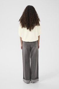 Sieviete ar gariem viļņotiem matiem, tērpusies vaļīgā baltā augšdaļā un platām pelēkām džinsa biksēm ar uzrollētām aprocēm, stāvot pretējā virzienā uz vienkārša fona.