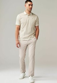 Beige mønstrede polo skjorte med korte ærmer, hvid krave, parret med lysebeige bukser og hvide sneakers. Enkel, casual design.