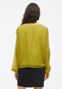 Limonengrüne, semi-transparente Bluse mit langen, gepufften Ärmeln und geradem Saum, kombiniert mit einem schwarzen Denim-Rock.