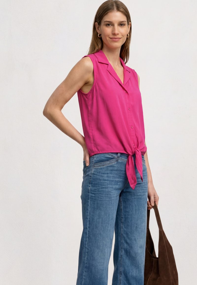 Femme portant un chemisier rose sans manches noué devant et un jean bleu, tenant un sac cabas marron, debout devant un fond clair uni.