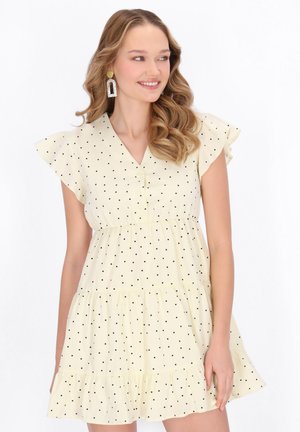 Fiatal nő hosszú, hullámos hajjal, világos sárga pöttyös rövid ujjú ruhában és nagy geometrikus fülbevalóval, mosolyogva oldalra néz.