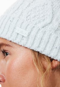 Lysegrå strikket beanie med kældesign, der har et struktureret ribbet mønster og små paljetter. Inkluderer et hvidt CMP-logo mærke.