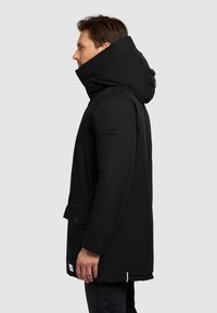 Czarna ocieplana parka z wysokim kołnierzem i regulowanym kapturem. Posiada duże przednie kieszenie, gładką teksturę oraz naszywkę z logo na rękawie.