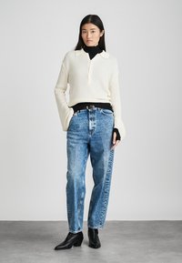 Suéter de punto color crema con cuello, cuello alto negro debajo, combinado con jeans de tiro alto de color azul claro y botas negras hasta el tobillo.