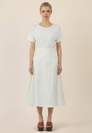 Frau steht mit dem Gesicht nach vorne, trägt ein weißes kurzärmeliges Oberteil mit Schulterauschnitt und einen hoch taillierten weißen Midirock sowie braune Plateauschuhe.