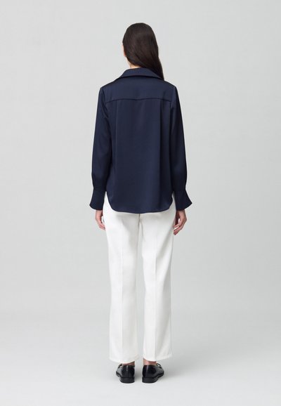 Camisa de satén azul marino de manga larga con collar clásico, corte relajado y puños con botones, combinada con pantalones amplios blancos.