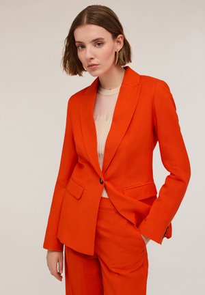 Heldere oranje, op maat gemaakte blazer met een enkele knoopsluiting, gestructureerde revers en twee voorzakken. Gedragen over een licht gestructureerd top.