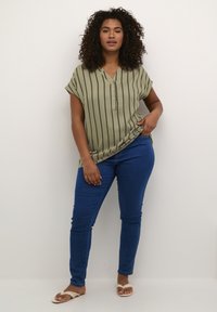Blouse vert olive à manches courtes avec des rayures verticales noires, patte de boutonnage, associée à un jean skinny bleu et des sandales légères.