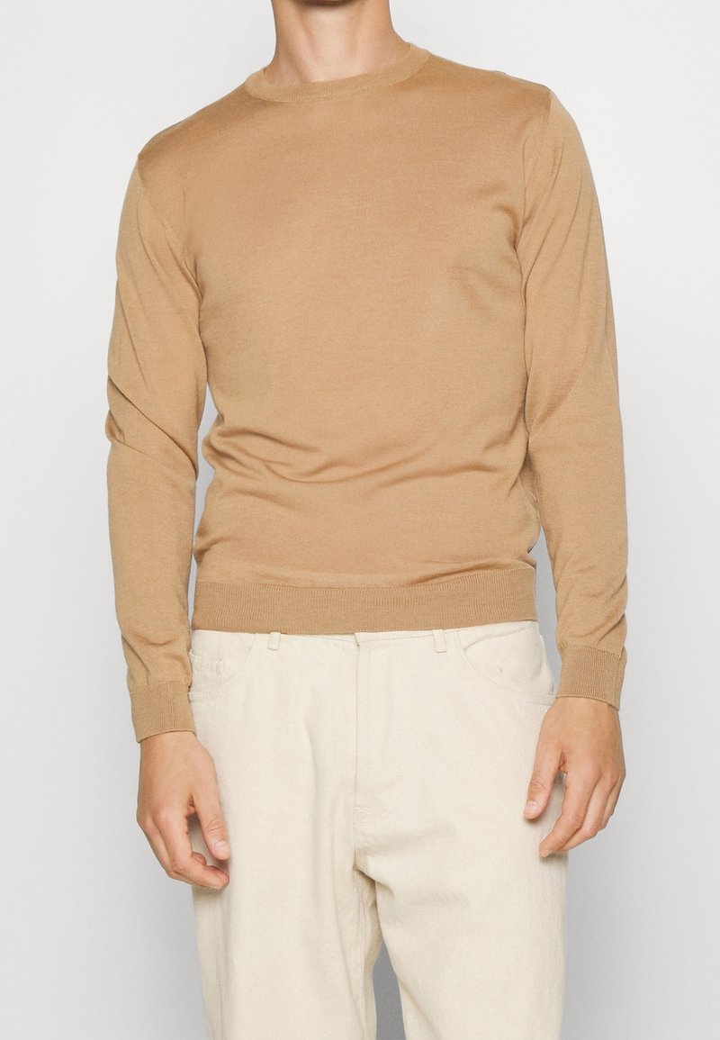 Homme portant un pull ajusté à manches longues couleur camel et un pantalon blanc cassé sur un fond blanc uni.