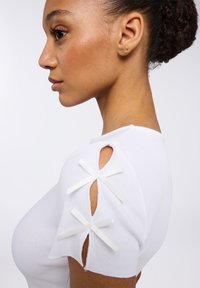 T-shirt bianco a righe con spalle scoperte decorate da fiocchi bianchi, design aderente e texture liscia.