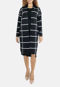 Long cardigan noir avec un motif à carreaux blancs, devant ouvert, deux poches, tissu tricoté texturé, longueur genou, porté sur une robe noire.