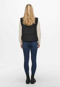 Chaleco tipo puffer negro con un diseño semicropped, llevado sobre un suéter de color claro, combinado con unos jeans ajustados azules y botas de tobillo oscuras.