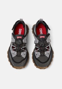 Czarno-szare buty outdoorowe mają teksturowaną podeszwę, elastyczne sznurowadła i czerwone logo. Wierzch buta ma akcenty z siateczki i zamszu dla lepszego wsparcia.
