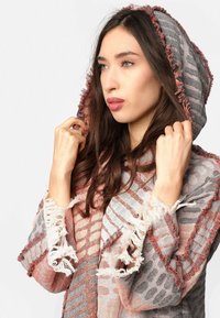 Kapucňový plášť z jemnej, textúrovanej tkaniny s šedými, ružovými a bielymi pruhmi. Obsahuje rozstrapkané okraje a zložitý vzor po celej ploche.