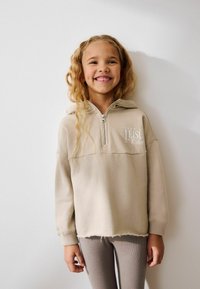 Beige zip-up hoodie med känguruficka, mjuk textur och en logotyp. Kombinerad med ljusbruna ribbade leggings, visad mot en neutral bakgrund.