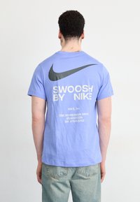 Ljuslila t-shirt i bomull, med en stor svart Nike swoosh-logotyp och vit text som säger "SWOOSH BY NIKE" på baksidan.
