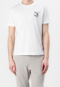 Hombre con una camiseta blanca con un pequeño gráfico de fruta cítrica negra en el pecho y pantalones gris claro frente a un fondo liso.