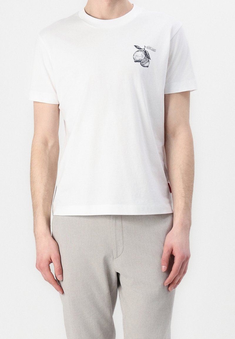 Hombre con una camiseta blanca con un pequeño gráfico de fruta cítrica negra en el pecho y pantalones gris claro frente a un fondo liso.