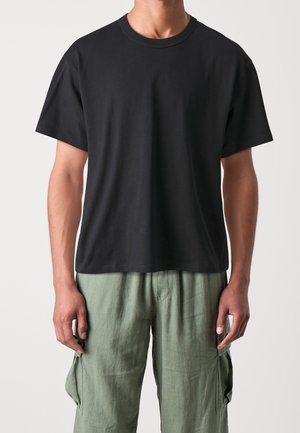 Manden iført en ensfarvet sort t-shirt med korte ærmer og lysegrønne cargo-bukser står mod en neutral baggrund.