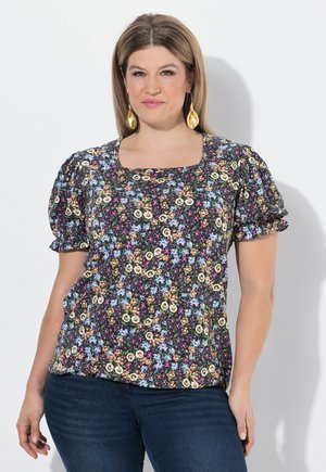 Frau mit schulterlangem Haar, die ein kurzärmeliges Blumenmuster-Oberteil und goldene Ohrhänger trägt, vor einem einfarbigen hellen Hintergrund stehend.